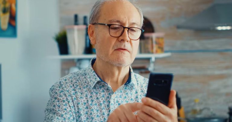 Un pensionar zâmbind în timp ce verifică o notificare de la Casa de Pensii pe ecranul telefonului său mobil